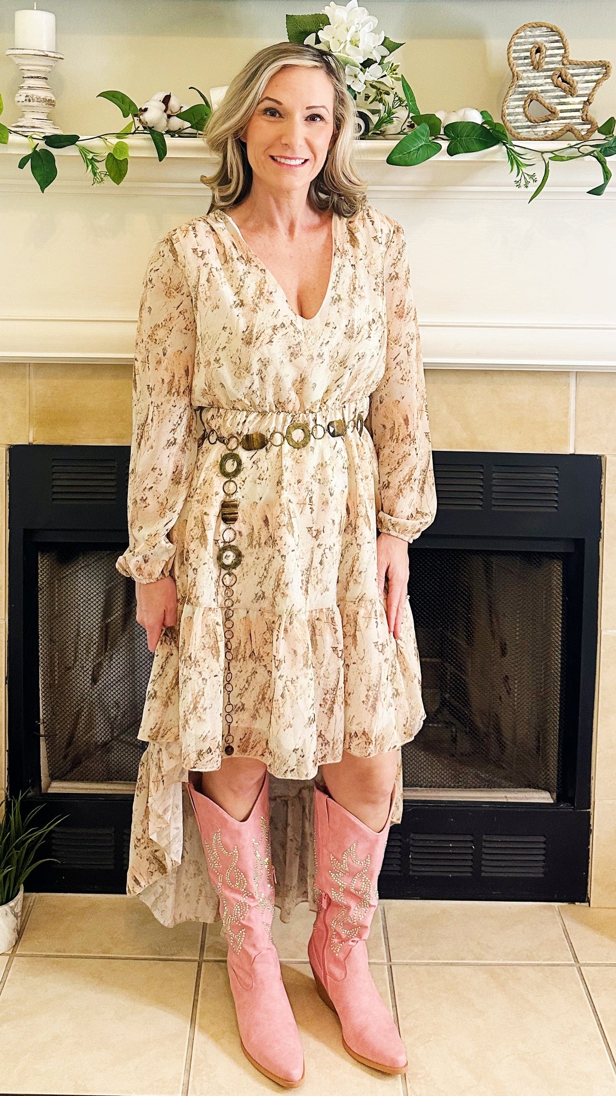 A woman stands in front of a fireplace wearing a long-sleeve beige floral dress with a buttoned waist and pink cowboy boots.
