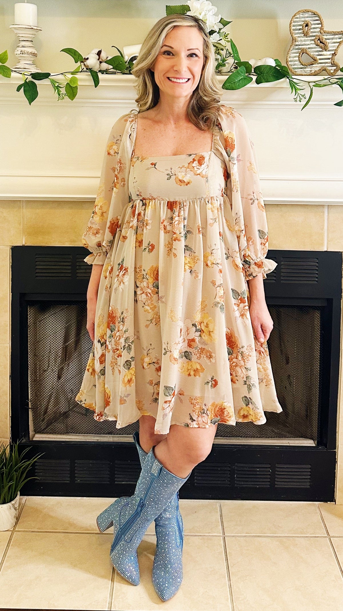 A woman stands in front of a fireplace wearing a beige floral dress with a sheer long-sleeve overlay and blue glittery knee-high socks or boot covers. The room has a cream fireplace mantel with greenery and decorative items.