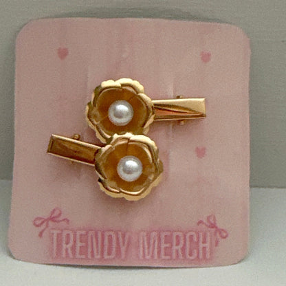A pair of gold-toned flower-shaped hair clips with white faux pearls in the center, displayed on a pink backing card that reads 'TRENDY MERCH'.