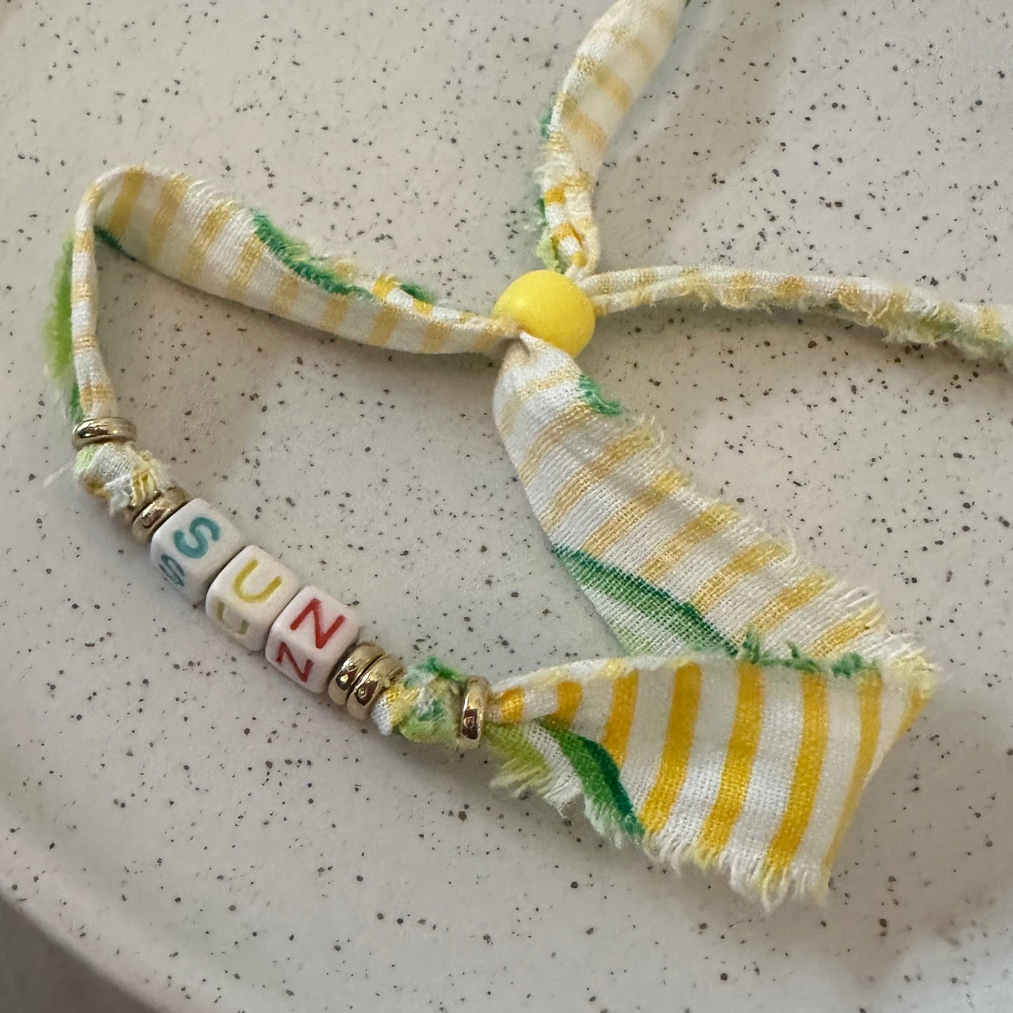 A handmade fabric bracelet or necklace made from yellow and white striped frayed ribbon, with a small yellow bead, gold spacer beads, and letter beads spelling 'SUN'.