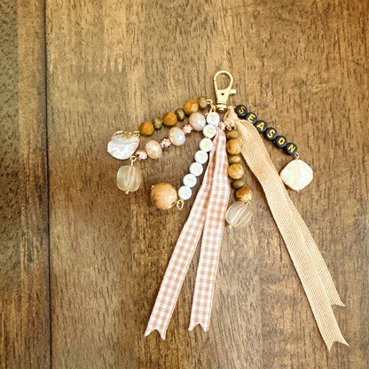 A wbohemian-style beaded keychain charm with wooden beads, clear glass beads, a gold clasp, a 'SEASON' bead string, and two hanging ribbons, displayed on a dark wood background.