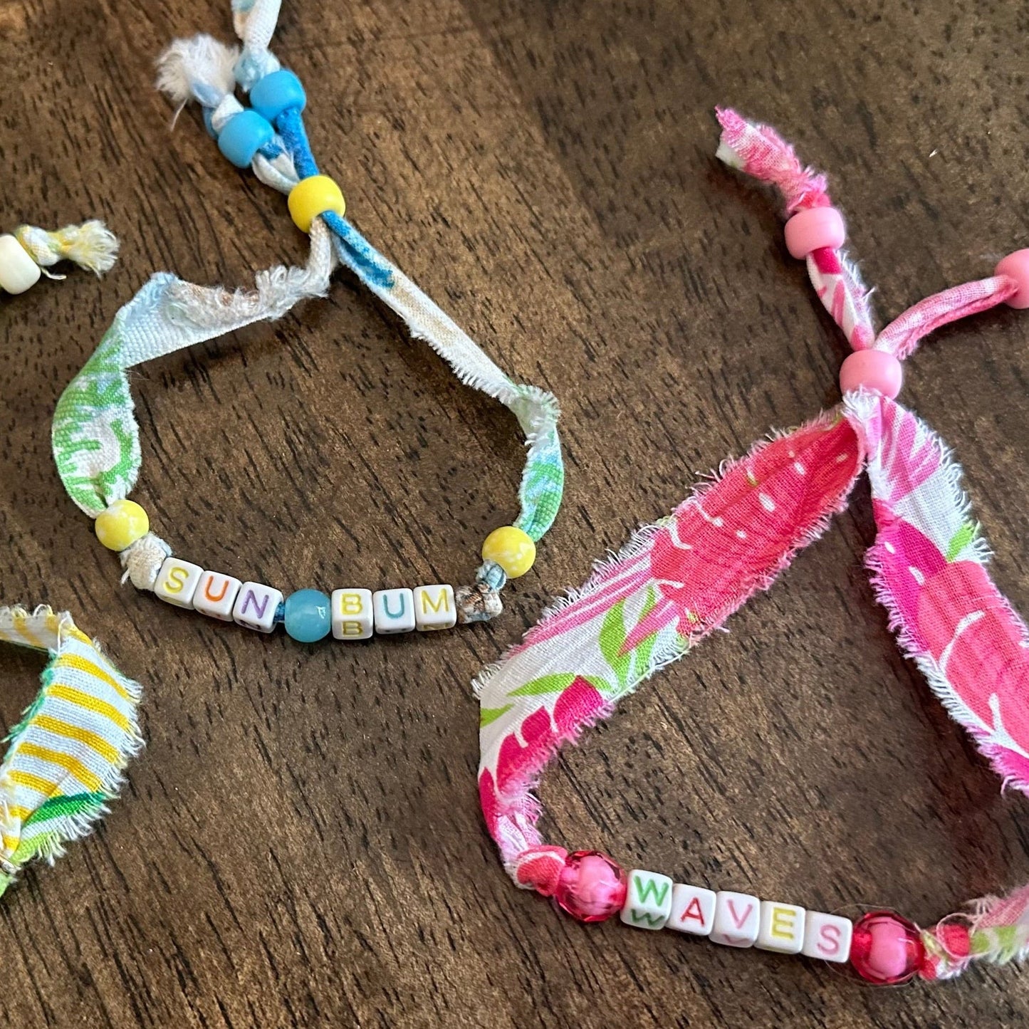Three handmade fabric beaded bracelets displayed on a dark wooden surface. The left bracelet is cream and green with yellow accents, the middle is green and white with blue  accents, and the right is pink and white with pink accents.