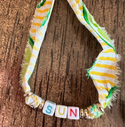 A handmade fabric necklace with frayed edges in yellow, white, and green stripes, featuring a yellow bead at the top and letter beads spelling 'SUN' on a dark wood background.