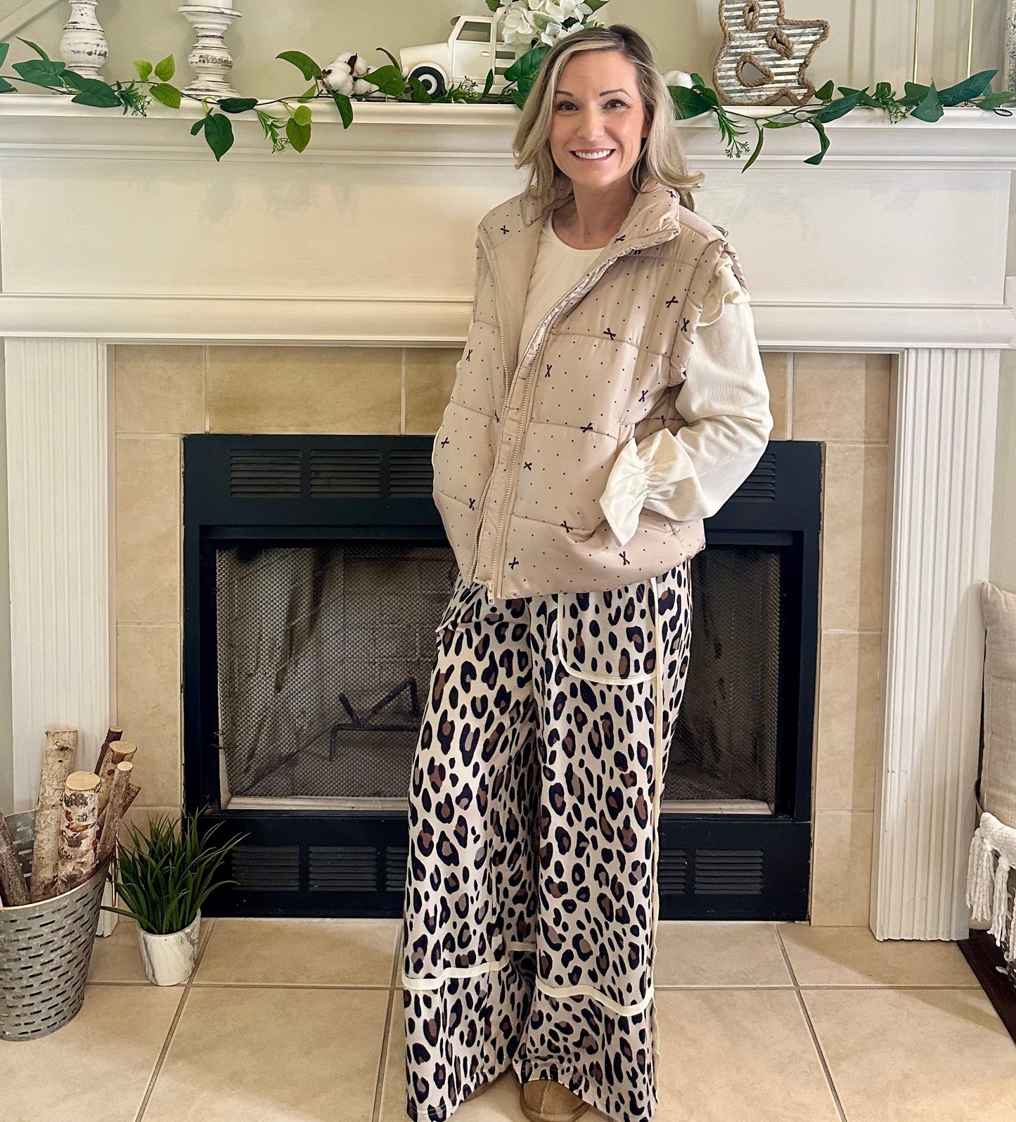 Woman standing in a living room with a fireplace, wearing a beige jacket and leopard print pants.