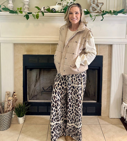 Woman standing in a living room with a fireplace, wearing a beige jacket and leopard print pants.
