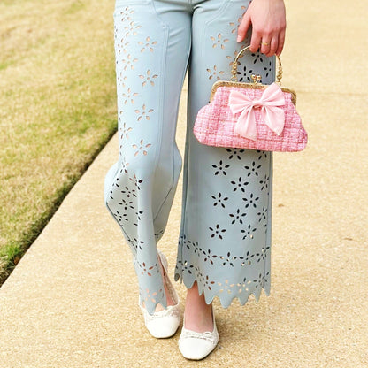 A person outdoors wearing light blue wide-leg pants with floral eyelet cutouts, a white blouse with pink ruffles, and holding a pink quilted handbag with a gold handle.
