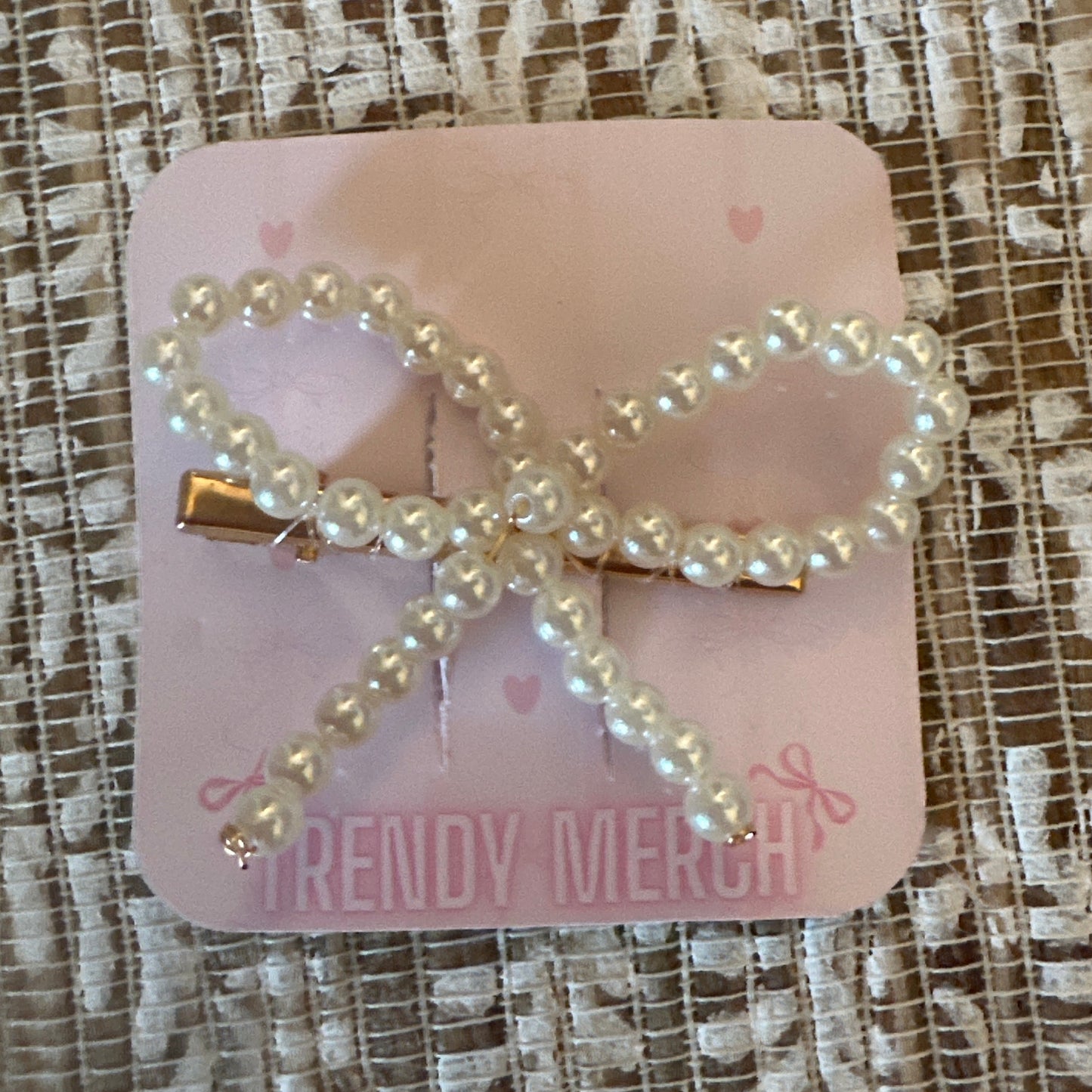 A pearl-bead bow hair clip with gold-tone metal hardware, attached to a pink display card that reads 'TRENDY MERCH', placed on a textured beige and white lace fabric background.