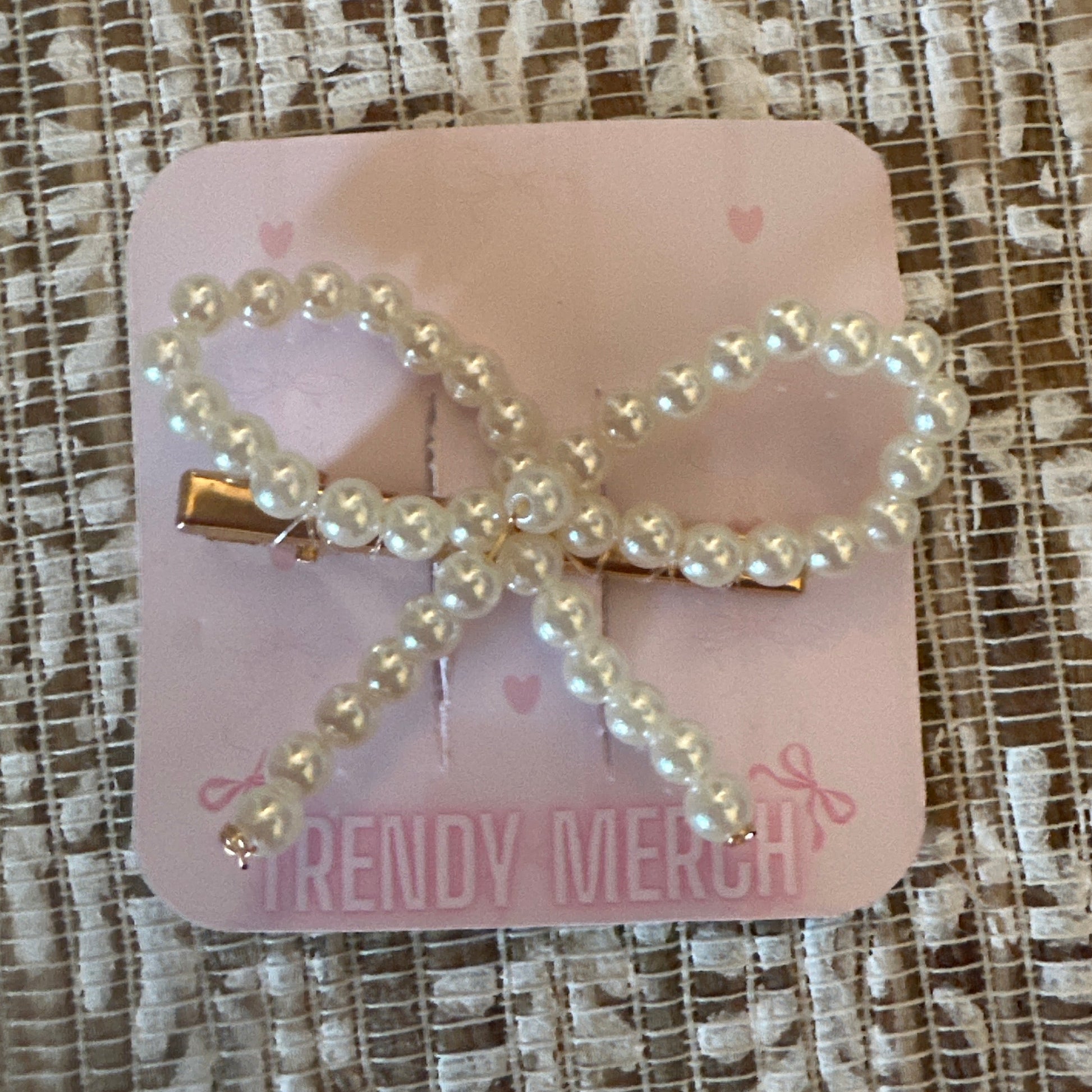 A pearl-bead bow hair clip with gold-tone metal hardware, attached to a pink display card that reads 'TRENDY MERCH', placed on a textured beige and white lace fabric background.