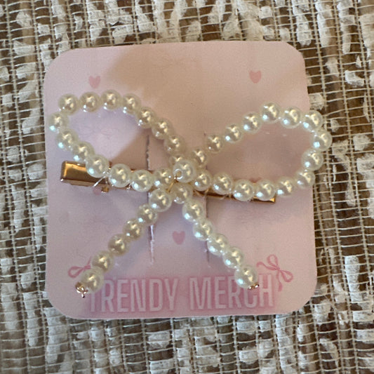 A pearl-bead bow hair clip with gold-tone metal hardware, attached to a pink display card that reads 'TRENDY MERCH', placed on a textured beige and white lace fabric background.