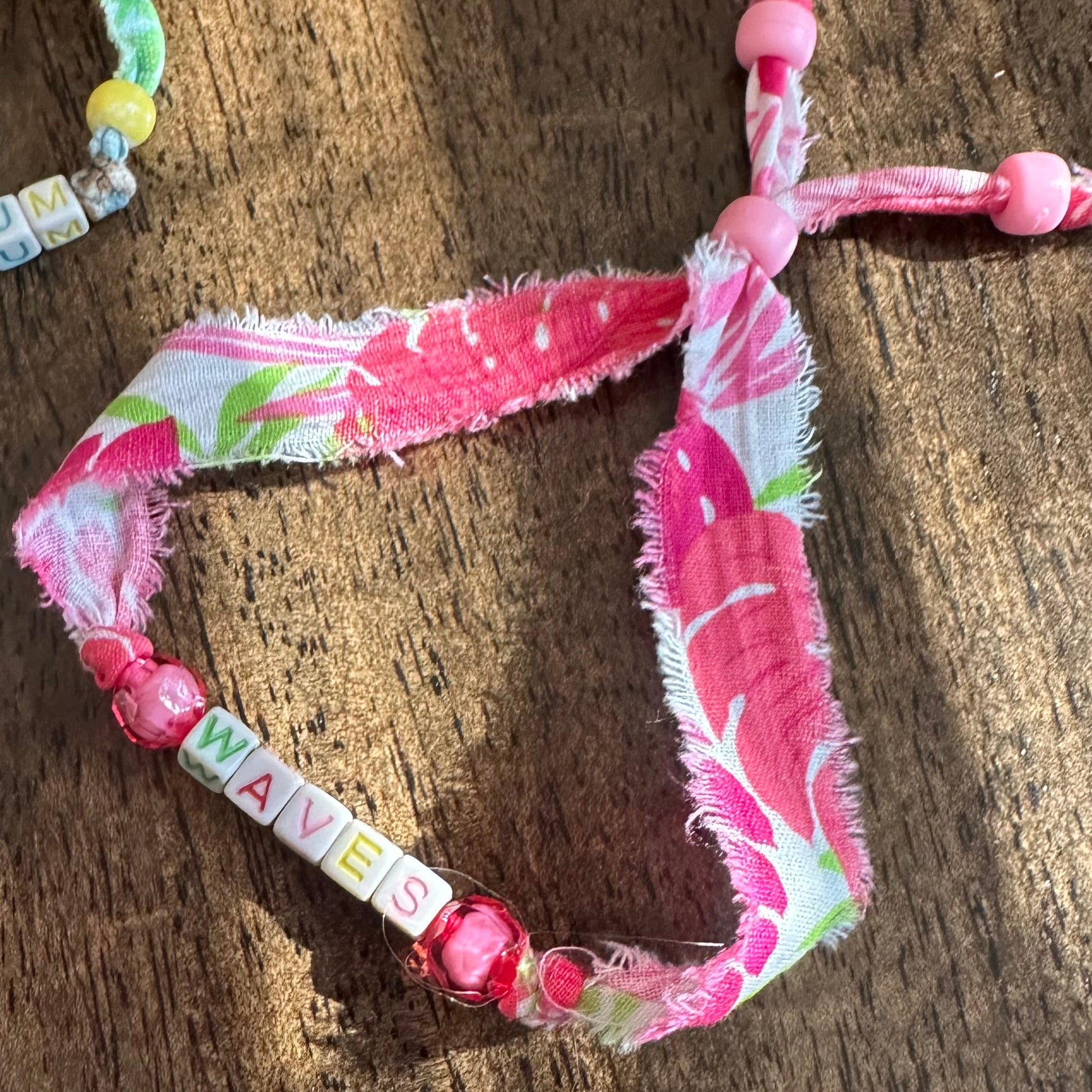 A handmade fabric bracelet with pink and white tropical floral print, frayed edges, pink beads, and a letter cube spelling 'NANAS', displayed on a wooden surface.