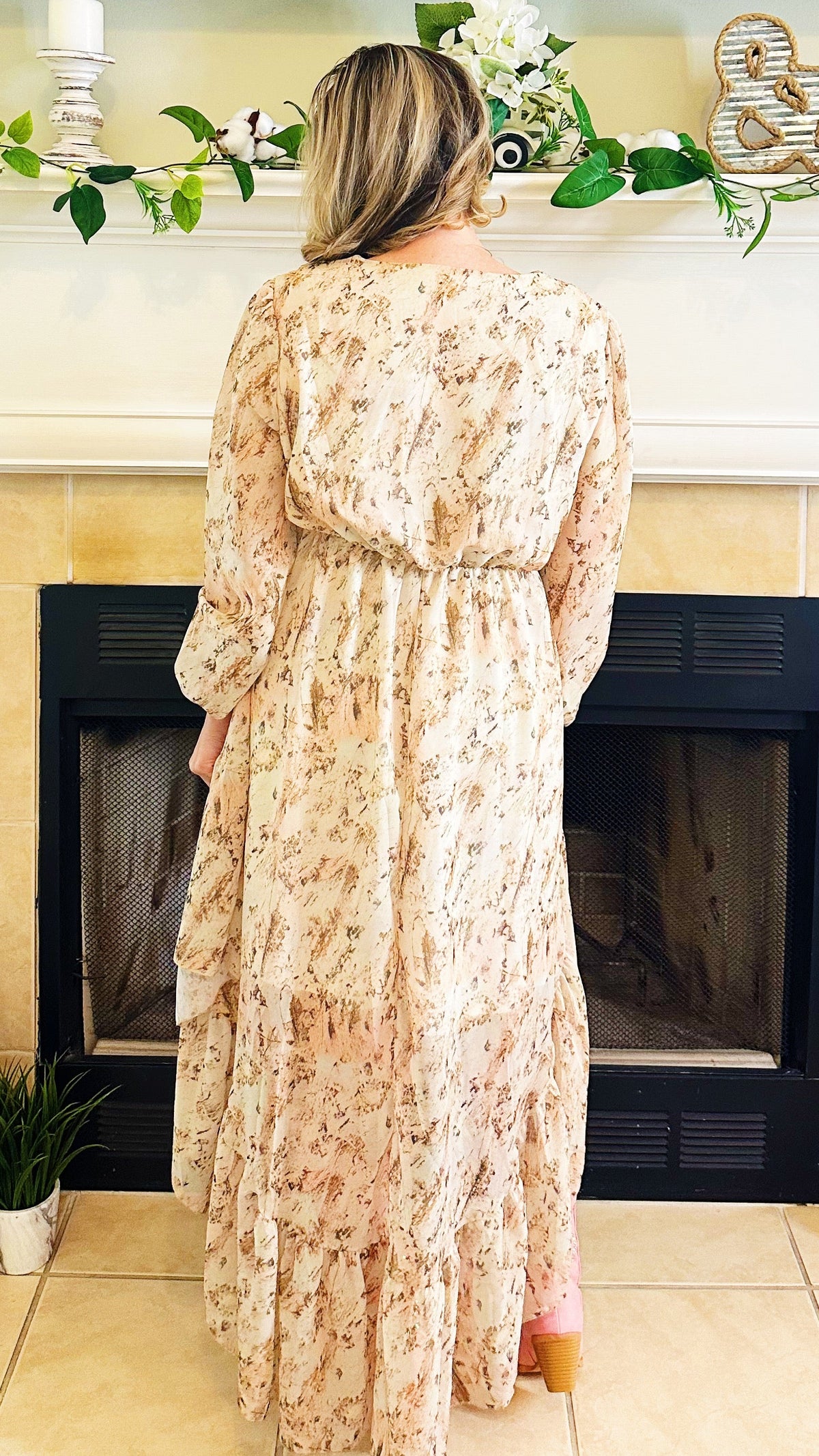 A woman viewed from behind wearing a long-sleeved, knee-length maxi dress with a beige and brown floral pattern, standing in front of a fireplace.