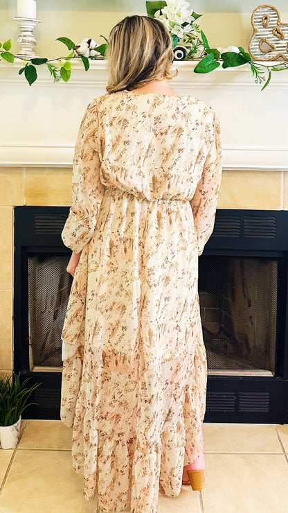 A woman viewed from behind wearing a long-sleeved, knee-length maxi dress with a beige and brown floral pattern, standing in front of a fireplace.