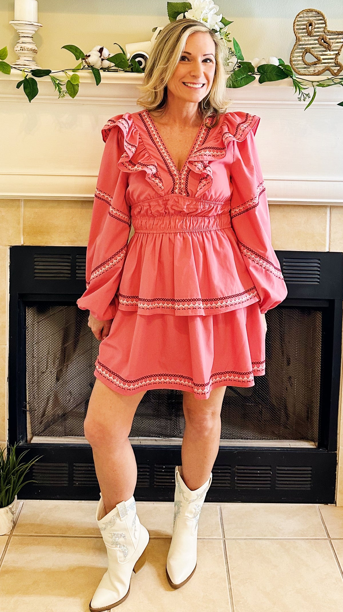 A woman wearing a coral pink tiered mini dress with ruffled sleeves and white cowboy boots, standing in front of a fireplace.