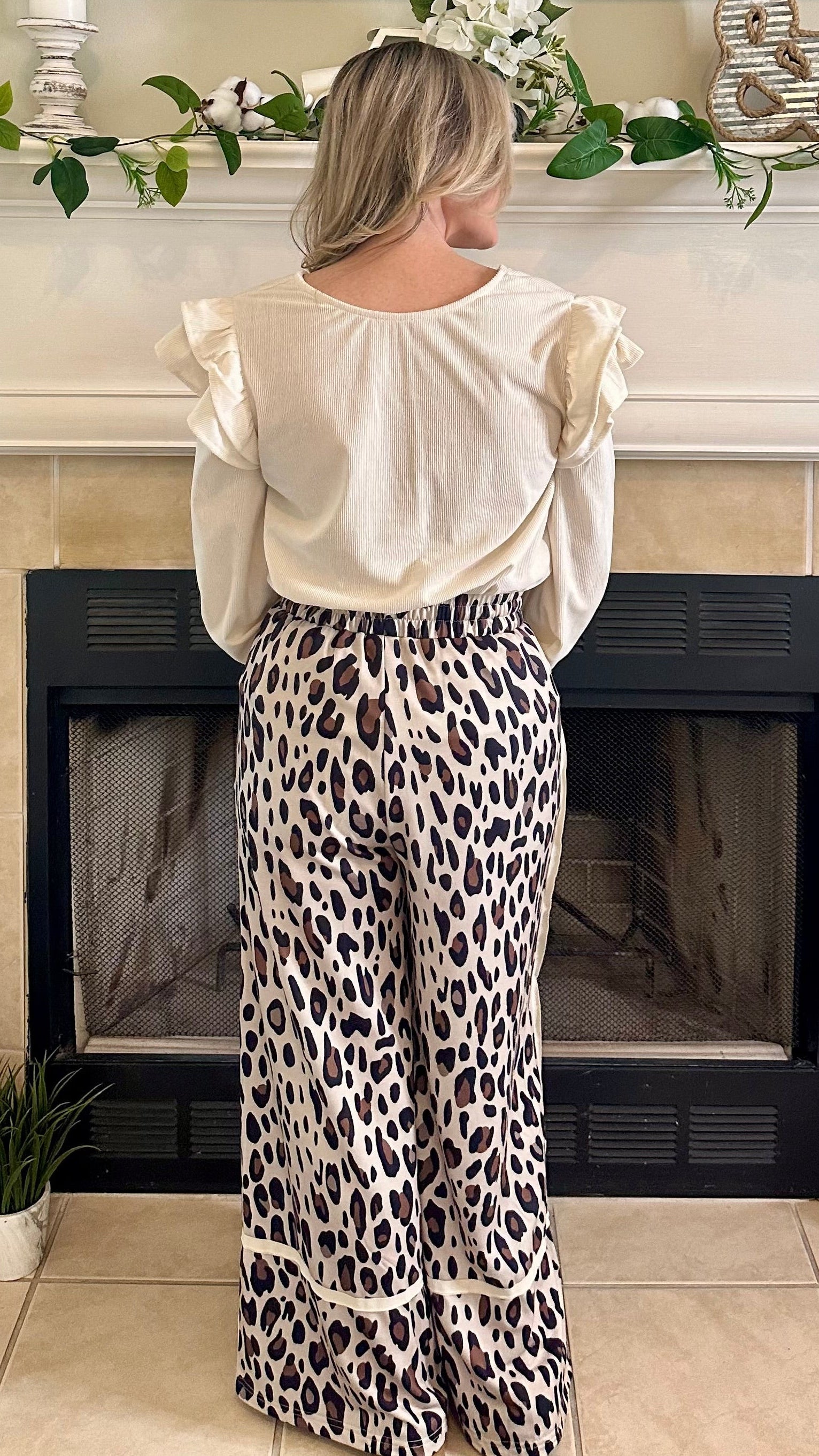 Woman in a white blouse and leopard print pants standing in front of a fireplace.