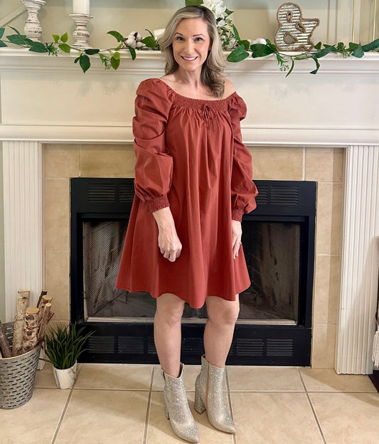 Woman in a rust color dress standing in front of a fireplace