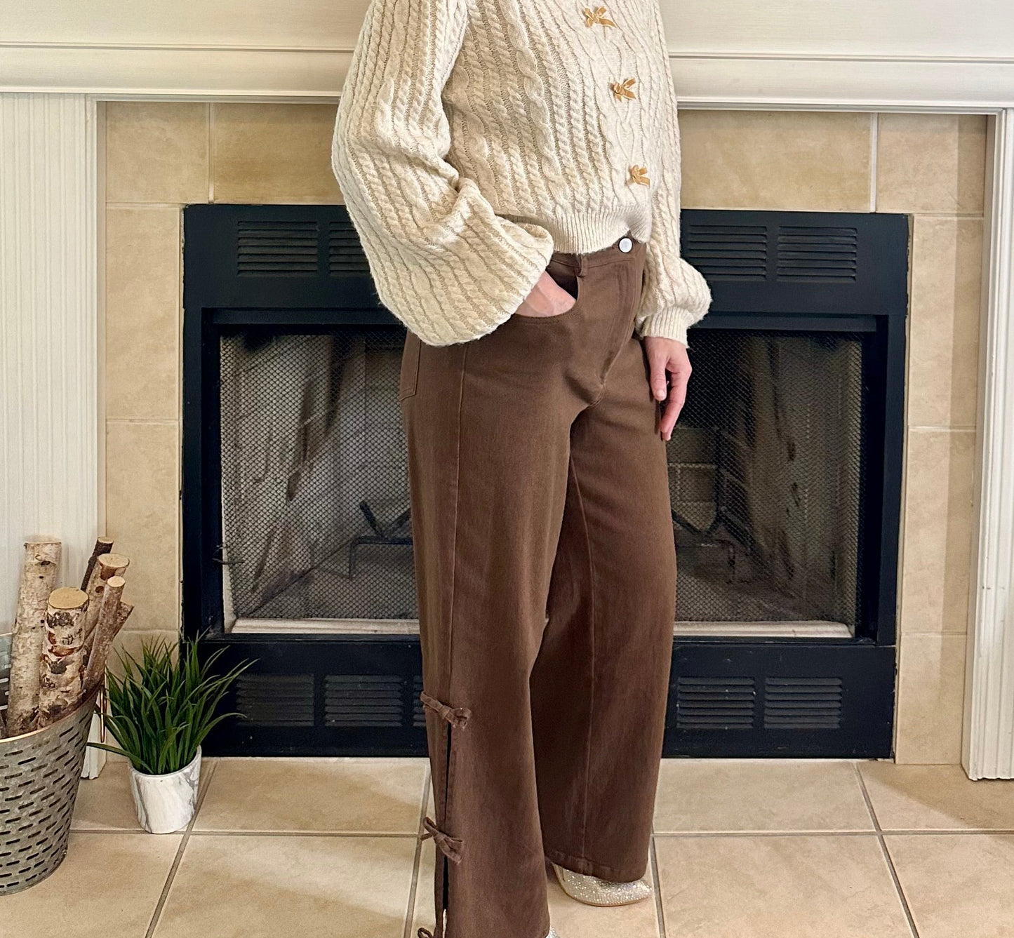 Woman standing in front of a fireplace wearing a cream sweater and brown pants.