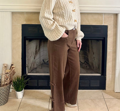 Woman standing in front of a fireplace wearing a cream sweater and brown pants.