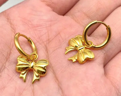 Gold bow-shaped earrings held in a hand against a neutral background