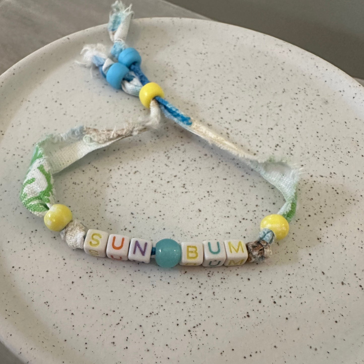 A handmade beaded bracelet with white fabric, yellow and blue beads, and letter tiles spelling 'SUN BUM' on a speckled white plate.
