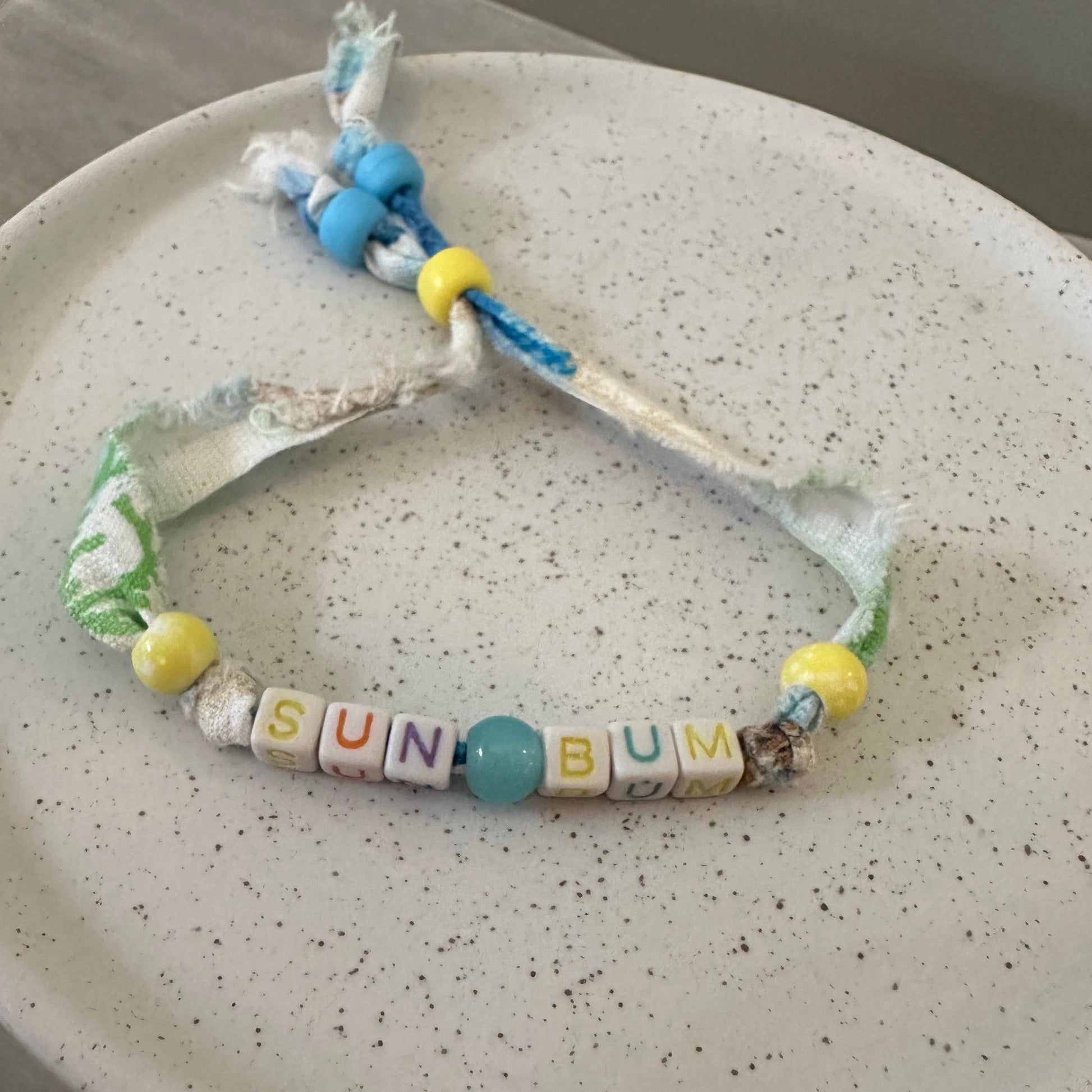 A handmade beaded bracelet with white fabric, yellow and blue beads, and letter tiles spelling 'SUN BUM' on a speckled white plate.