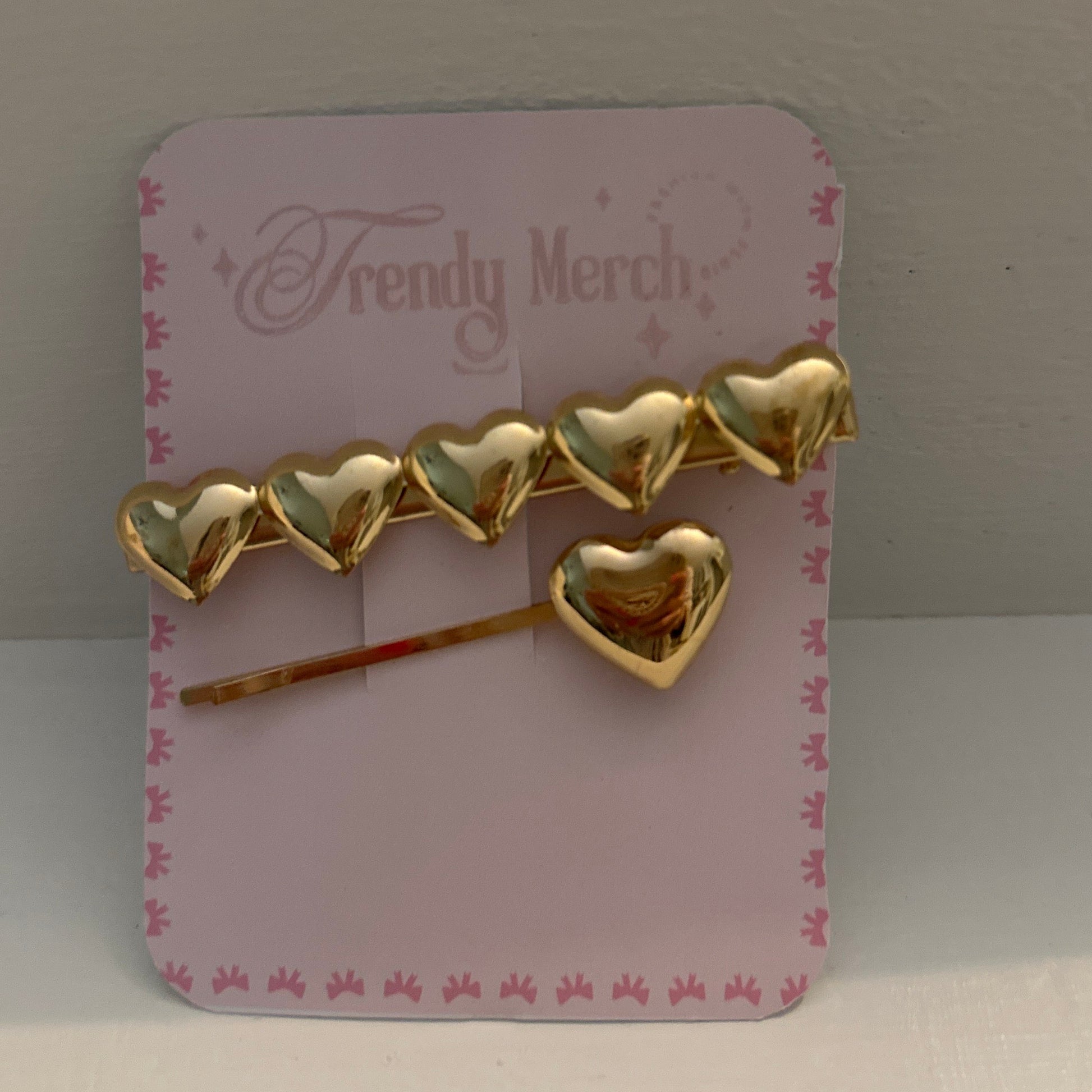 A gold heart-shaped hair accessory set displayed on a pink card labeled 'Trendy Merch,' photographed against a plain light background.