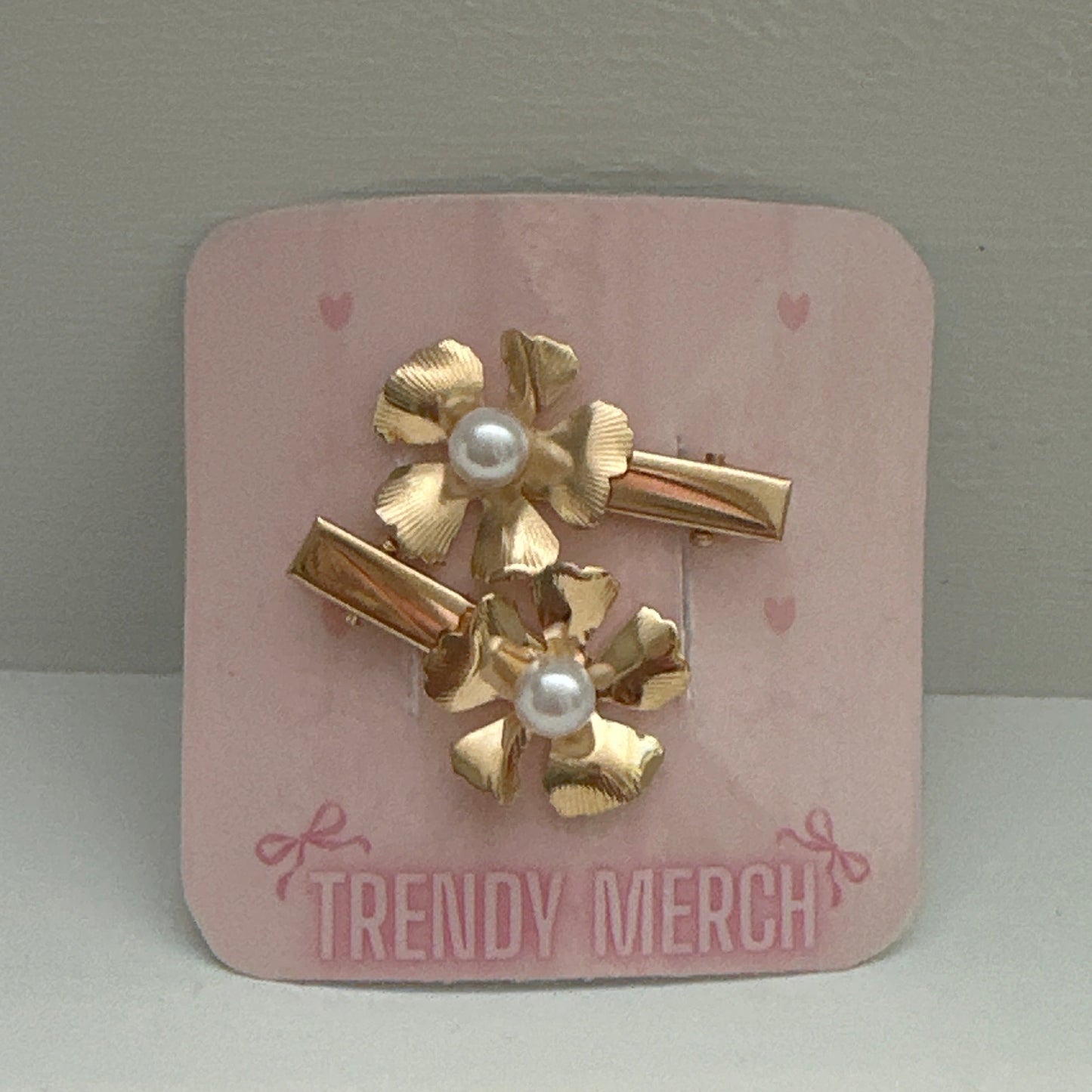 A set of three gold-tone hair clips with textured flower petals and white faux pearls, attached to a pink display card labeled 'TRENDY MERCH' against a neutral gray background.