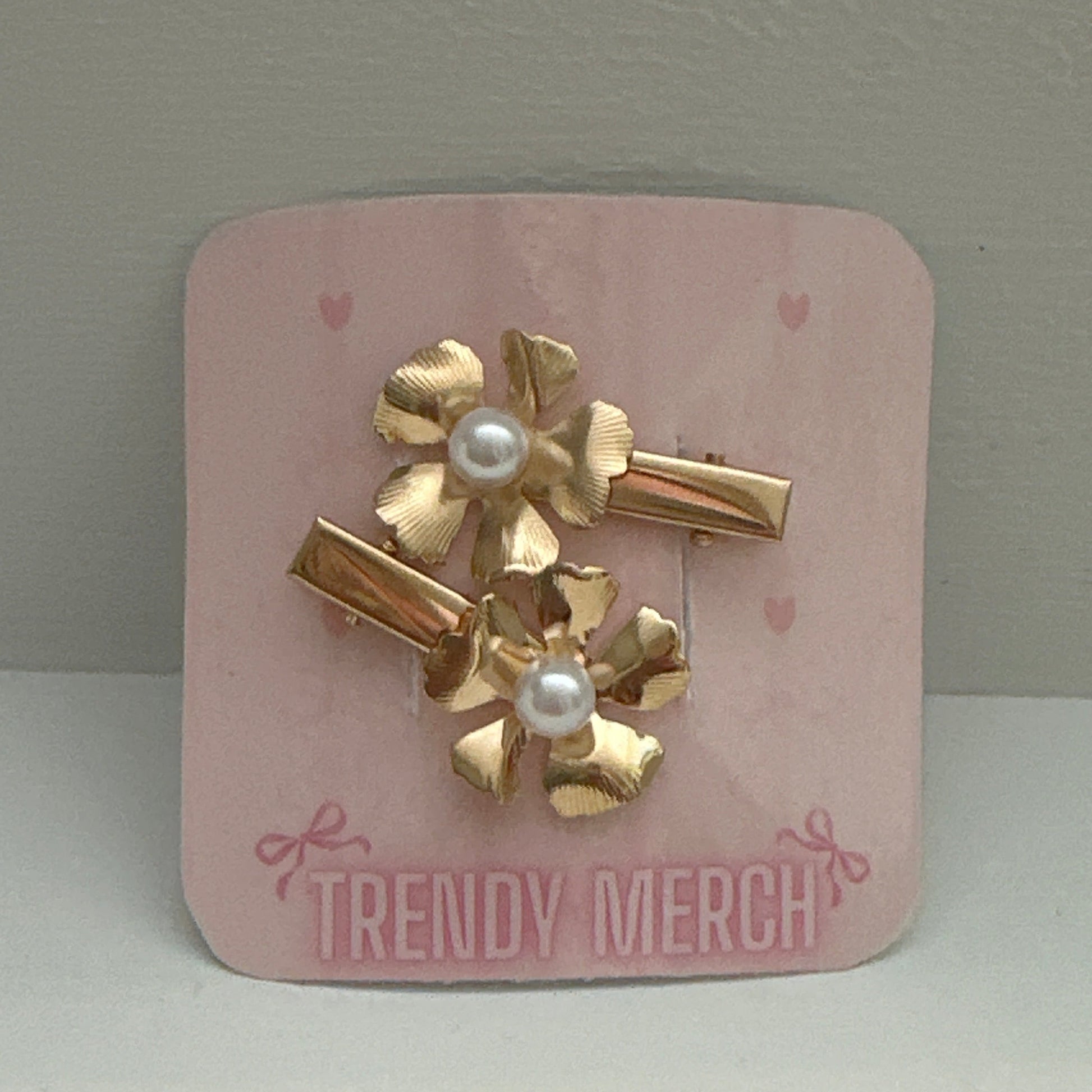 A set of three gold-tone hair clips with textured flower petals and white faux pearls, attached to a pink display card labeled 'TRENDY MERCH' against a neutral gray background.