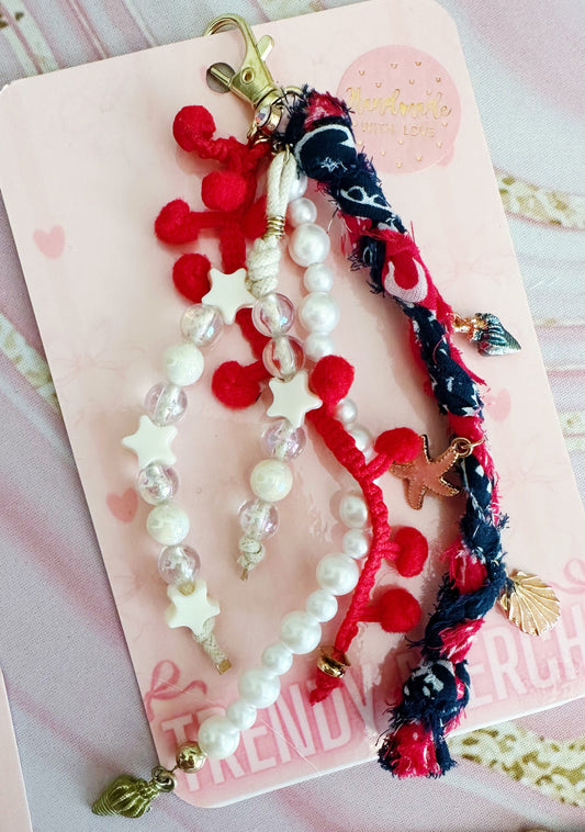 A set of nautical-themed charm keychains on a pink backing card. The keychains include white faux pearls with star-shaped beads, red pom-poms, clear beads, a braided navy and red cord with a shell charm, and a small gold anchor charm.