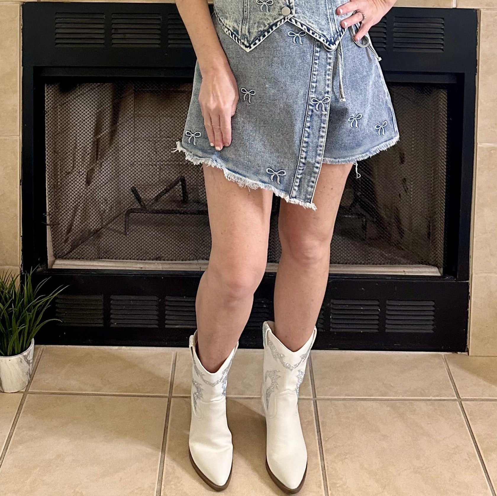 Woman in a denim dress and white boots standing in front of a fireplace.