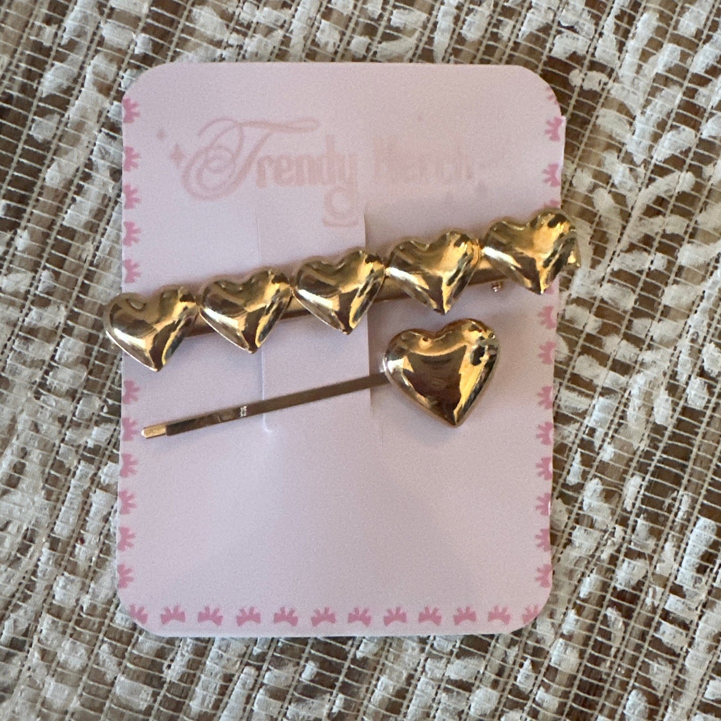 A gold heart-shaped hair accessory attached to a pink display card, placed on a textured beige lace fabric background. The card has the brand name 'Frendy Fierce' printed on it.