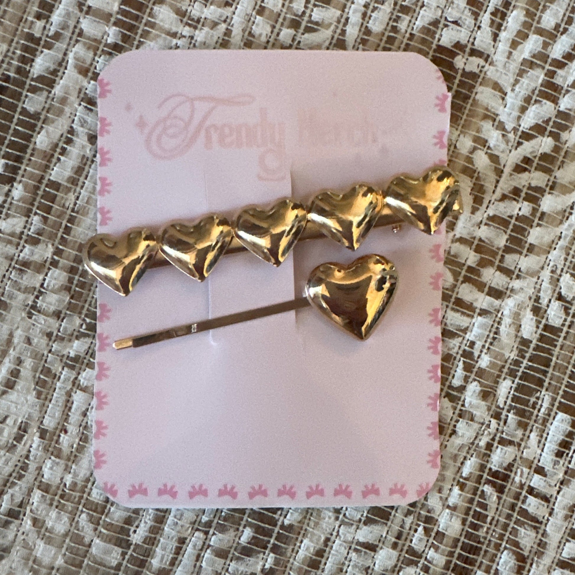 A gold heart-shaped hair accessory attached to a pink display card, placed on a textured beige lace fabric background. The card has the brand name 'Frendy Fierce' printed on it.