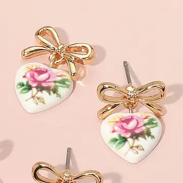 Heart-shaped earrings with floral design and gold bows on a pink background