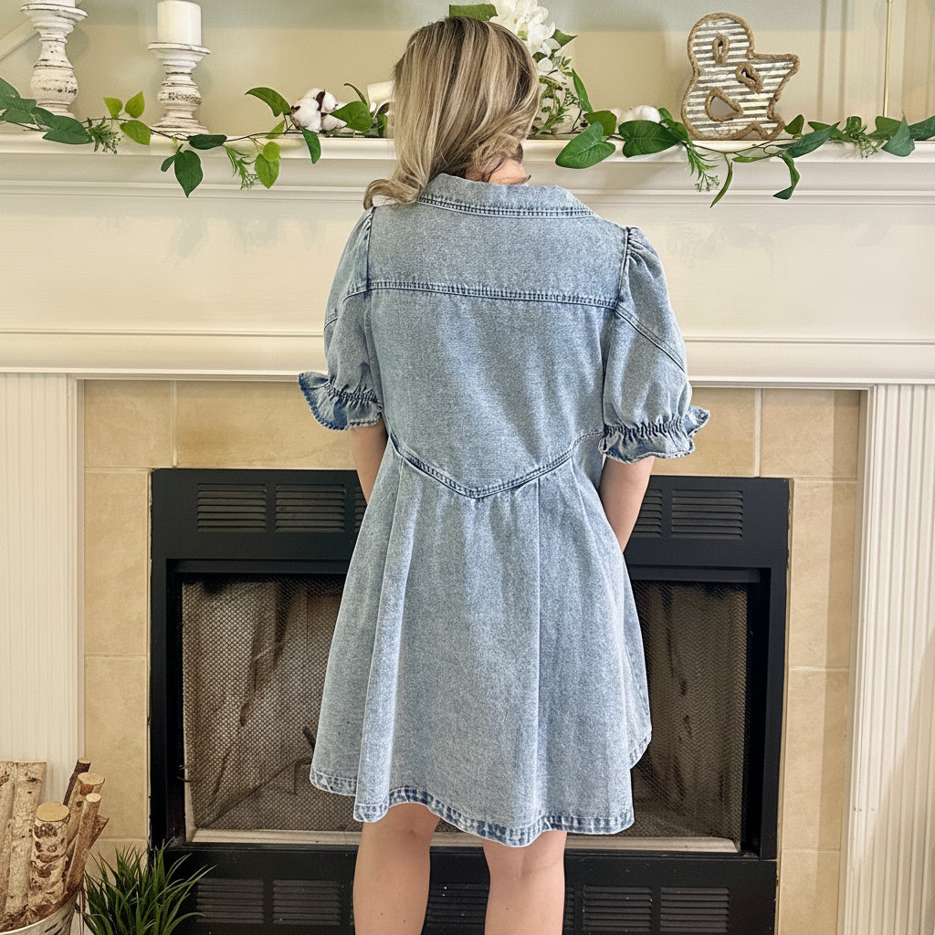 Woman in a denim dress standing in front of a fireplace with decorative items on the mantel.