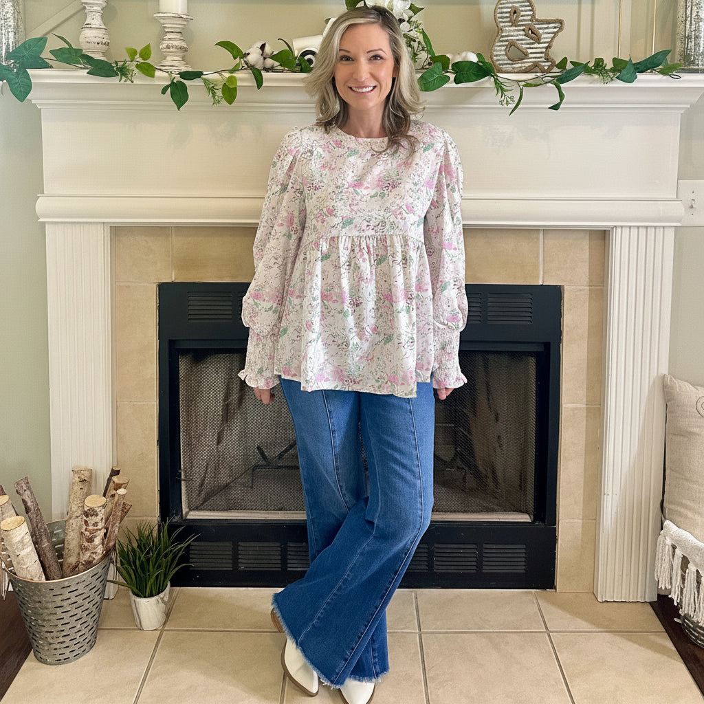 Woman standing in a living room with a fireplace, wearing a floral blouse and blue jeans.