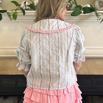 Person wearing a white blouse with pink ruffles and a pink skirt in front of a fireplace.