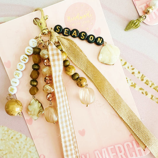 A wbohemian-style beaded keychain charm with wooden beads, clear glass beads, a gold clasp, a 'SEASON' bead string, and two hanging ribbons, displayed on a dark wood background.