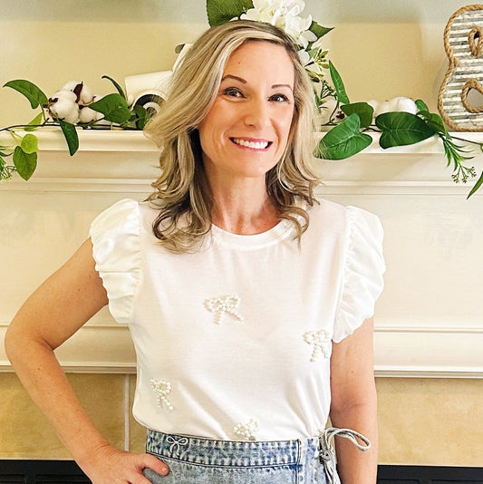 A woman posing in front of a fireplace wearing a white ruffle-sleeve top, a light-wash denim wrap mini skirt, and white western-style boots.