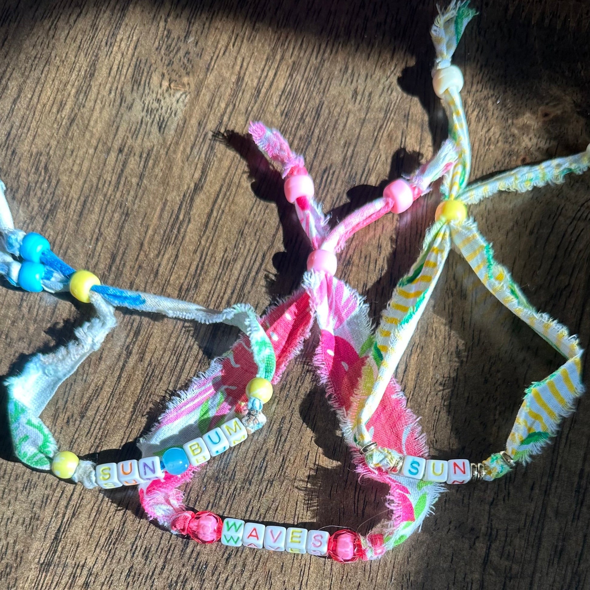 Three colorful fabric friendship bracelets with beads and letter tiles, laid out on a wooden surface in direct sunlight.