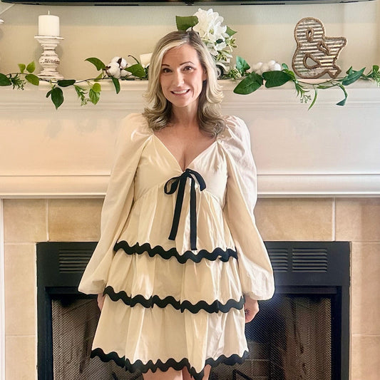 Woman in a beige dress with black trim standing in front of a fireplace.
