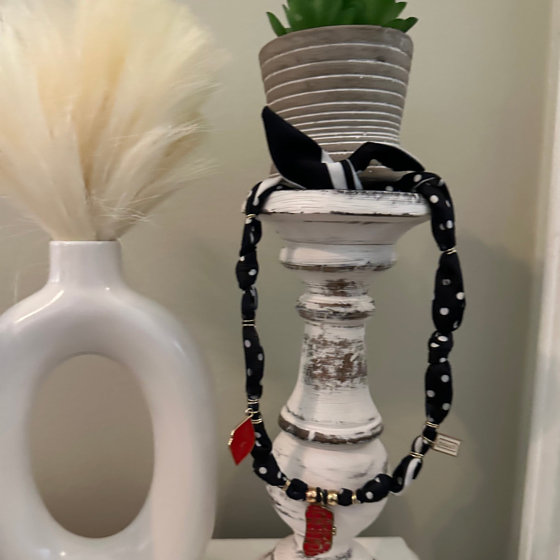 A white distressed wooden candlestick holder with a black and white polka dot chain leash and small red leather accents, shown next to a white toilet brush in a white ceramic holder.