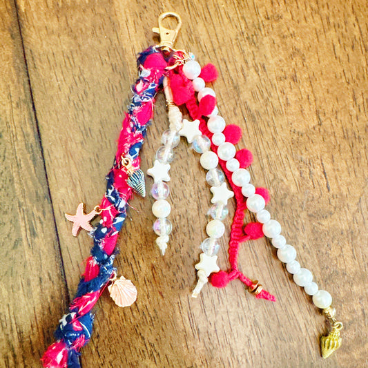A handmade keychain or bag charm featuring multiple fabric and bead strands on a dark wood background. The design includes a navy and red patterned ribbon, red pom-poms, white pearl-like beads, white star charms, a pink starfish, a seashell charm, and a gold lobster clasp.