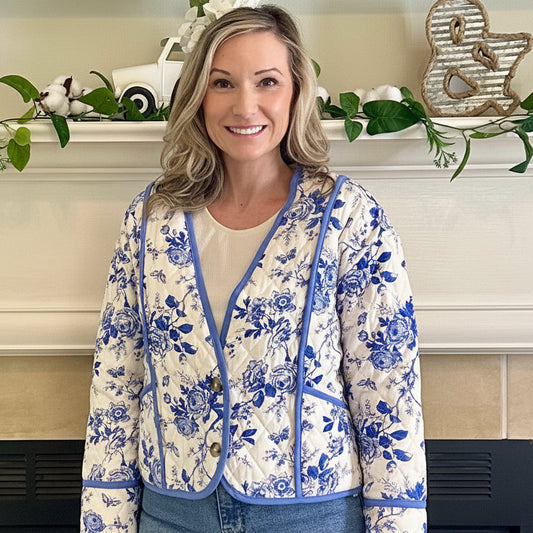 Woman wearing a floral jacket and jeans standing in front of a fireplace.
