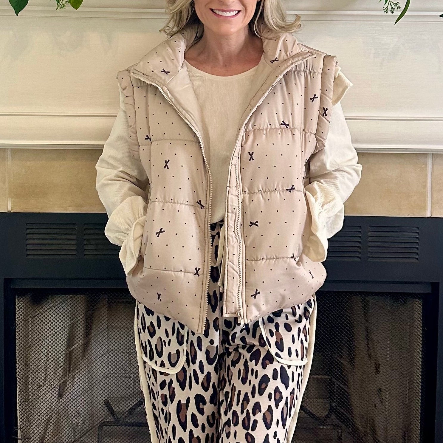 Person wearing a beige puffer vest over a light-colored top and leopard print pants, standing in front of a fireplace.