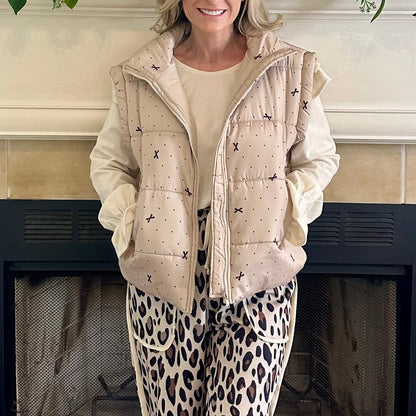 Person wearing a beige puffer vest over a light-colored top and leopard print pants, standing in front of a fireplace.