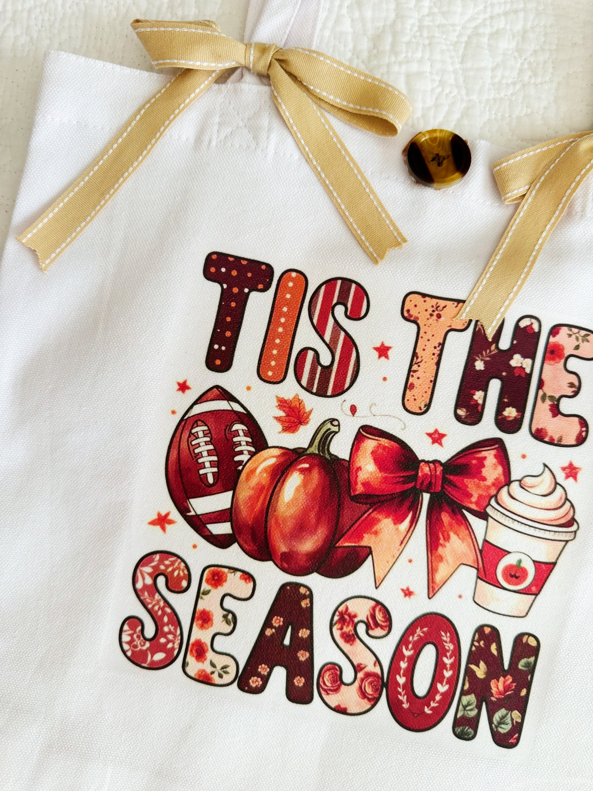 A white canvas tote bag with a gold ribbon handle bow, featuring a fall-themed graphic that reads 'TIS THE SEASON' in autumn patterned letters, alongside pumpkins, a football, a bow, and a hot cocoa cup.