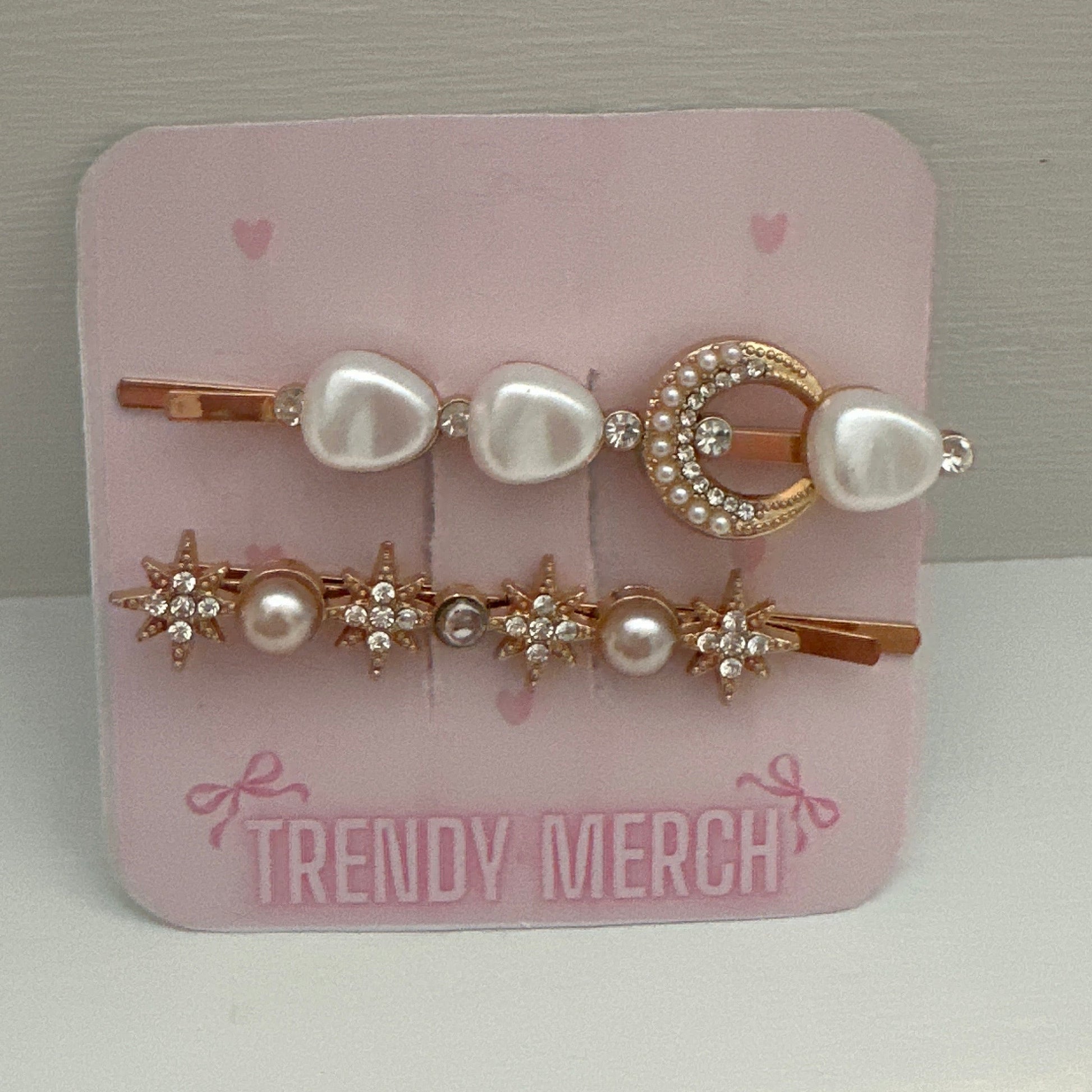 A set of gold  hair clips on a pink display card labeled 'TRENDY MERCH'. The clips feature white faux pearls and rose gold metal details, including a crescent moon and star designs, set against a neutral gray background.