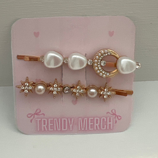 A set of gold  hair clips on a pink display card labeled 'TRENDY MERCH'. The clips feature white faux pearls and rose gold metal details, including a crescent moon and star designs, set against a neutral gray background.