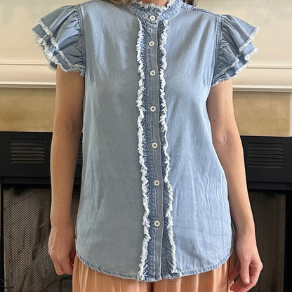 Person wearing a light blue denim shirt with ruffled details in front of a fireplace.