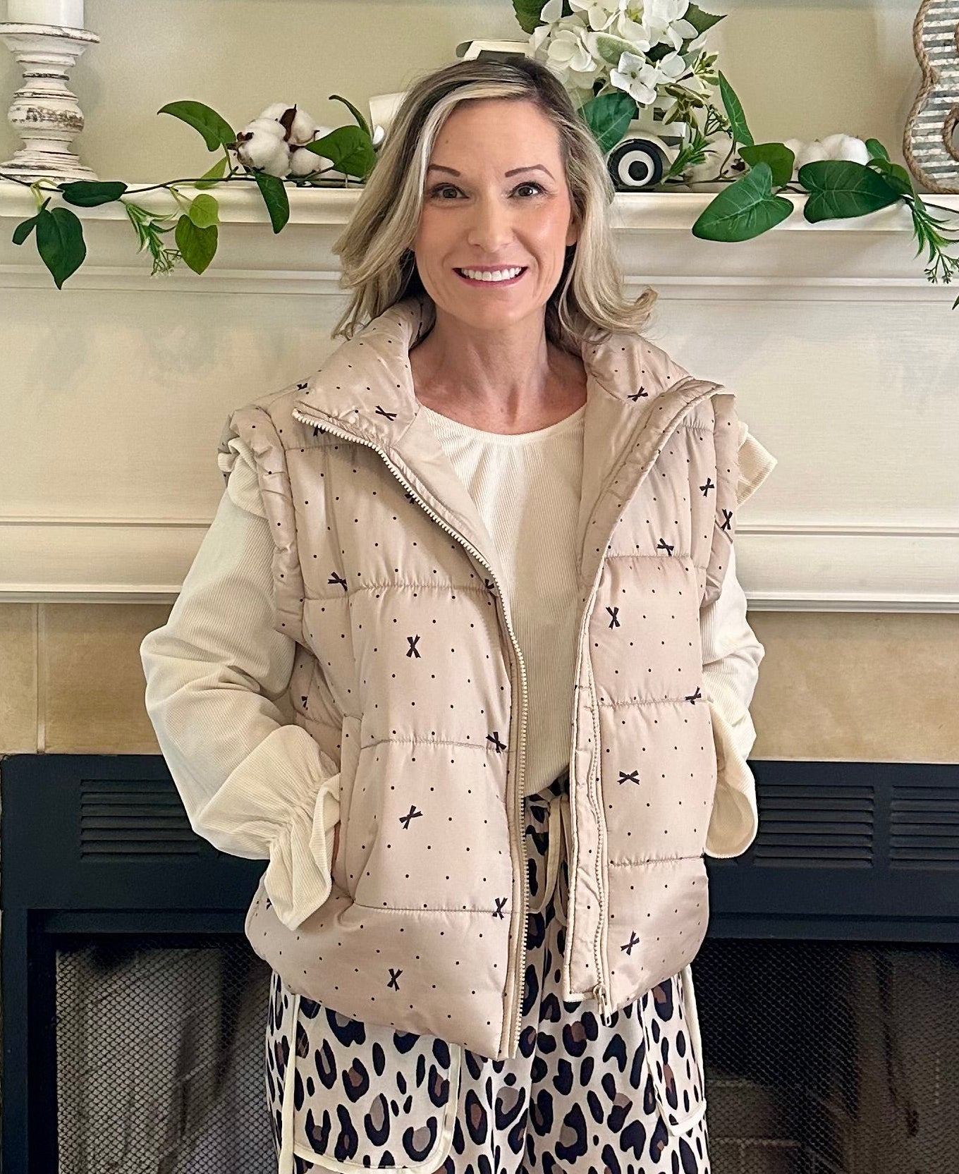 Woman standing in a living room with a fireplace, wearing a beige puffer vest and leopard print pants.