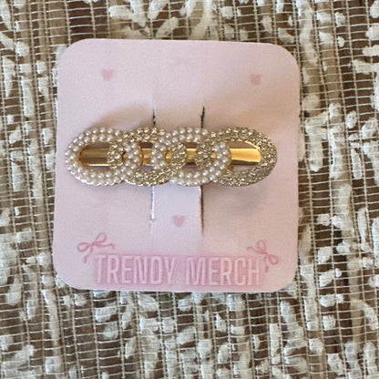 A gold-tone hair claw clip embellished with clear rhinestones, displayed on a pink card labeled 'TRENDY MERCH' against a textured beige and white lace fabric background.